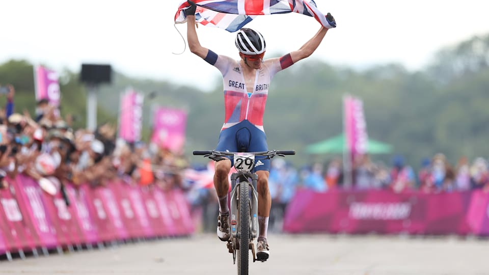 Thomas Pidcock celebrates winning the gold medal while holding the flag of his country during the Men's Cross-country race at Tokyo 2020.
